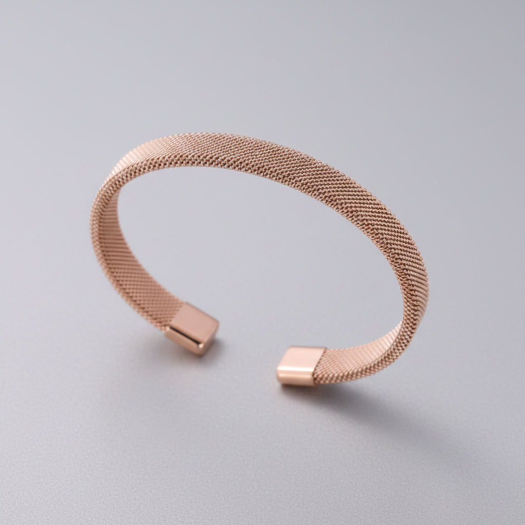 A slim open cuff bracelet with a fine mesh-like metallic texture in a warm rose gold tone, resting on soft draped fabric.