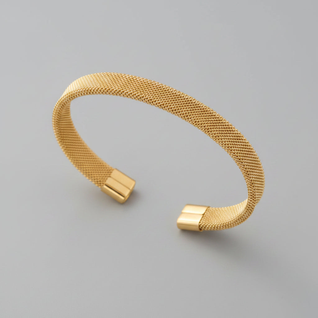 A gold-toned open cuff bracelet with a fine mesh-like woven texture, displayed on soft draped beige fabric.